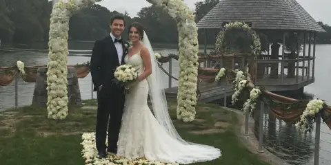 A bride and groom standing under a large floral arch with a flower-lined aisle at a waterfront venue.