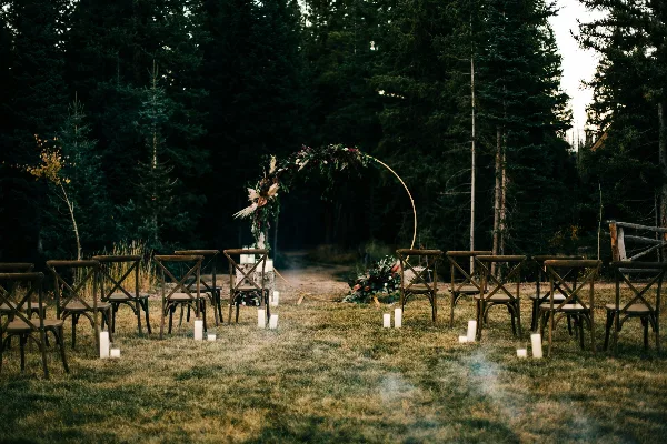 An outdoor wedding ceremony setup featuring a floral arch and rows of wooden chairs set on a grassy lawn surrounded by tall evergreen trees.