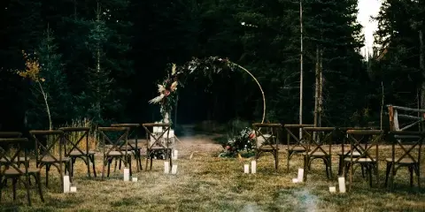 An outdoor wedding ceremony setup featuring a floral arch and rows of wooden chairs set on a grassy lawn surrounded by tall evergreen trees.
