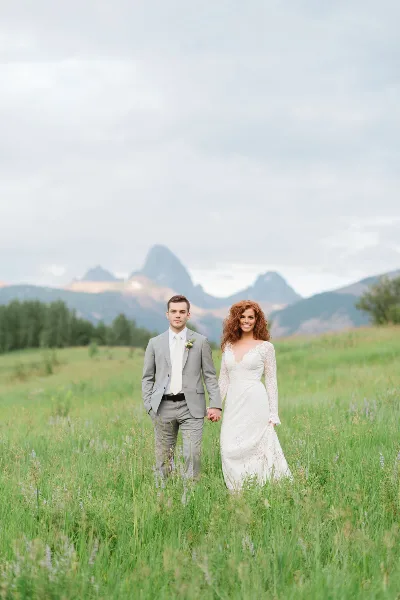 A bride and groom standing in a grassy field with mountains in the background.