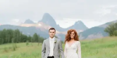 A bride and groom standing in a grassy field with mountains in the background.