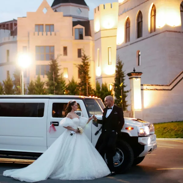 A bride and groom standing next to a white limousine in front of a castle-like building.