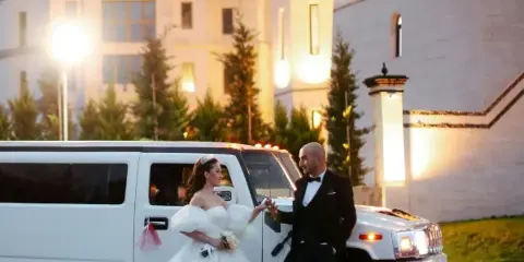 A bride and groom standing next to a white limousine in front of a castle-like building.