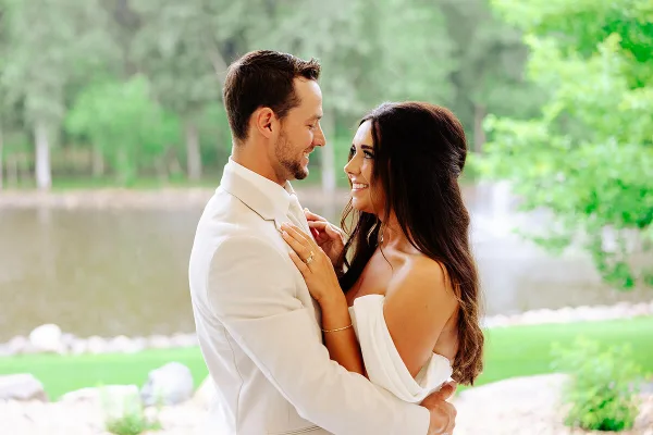 A smiling couple in wedding attire embracing outdoors near a lake.
