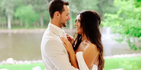 A smiling couple in wedding attire embracing outdoors near a lake.