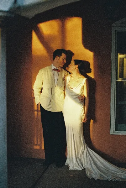 A wedding couple in formal attire standing together in a warm, sunlit doorway.