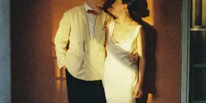 A wedding couple in formal attire standing together in a warm, sunlit doorway.