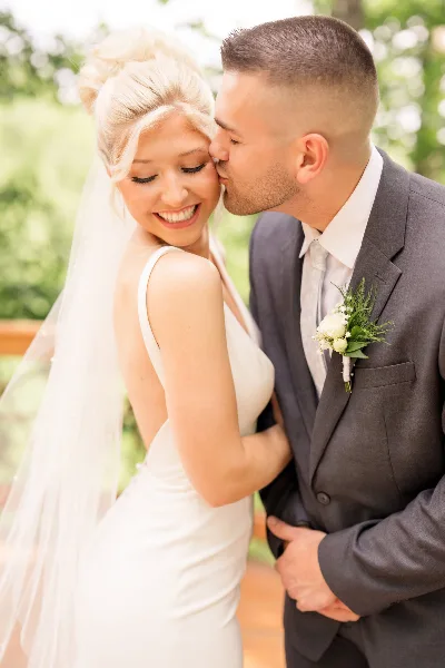 A bride in a white gown and veil smiling as a groom in a grey suit kisses her forehead.
