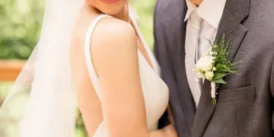 A bride in a white gown and veil smiling as a groom in a grey suit kisses her forehead.