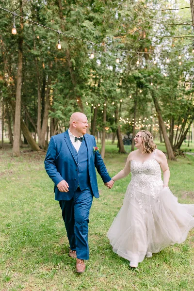 Bride and groom walking hand-in-hand on a grassy lawn under string lights in a wooded outdoor wedding venue setting.