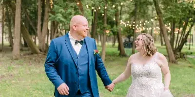 Bride and groom walking hand-in-hand on a grassy lawn under string lights in a wooded outdoor wedding venue setting.