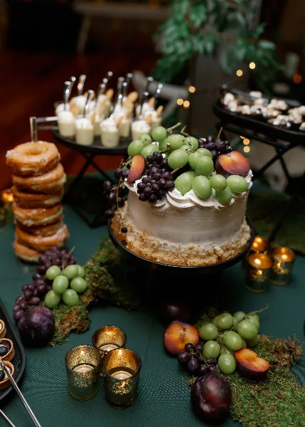 Wedding dessert table with a fruit-topped cake, pastries, desserts, grapes, and candlelight.