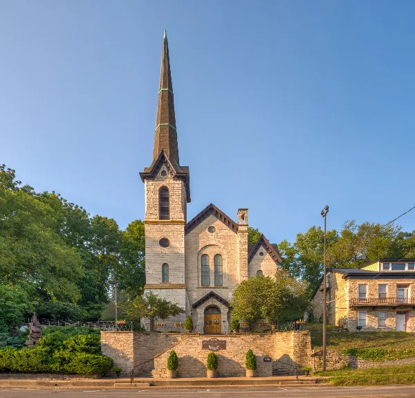 Exterior view of a historic stone church building with a tall steeple and surrounding greenery.