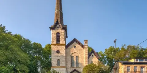 Exterior view of a historic stone church building with a tall steeple and surrounding greenery.