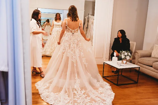 A bride trying on a lace wedding gown in a bridal boutique fitting room with a consultant.