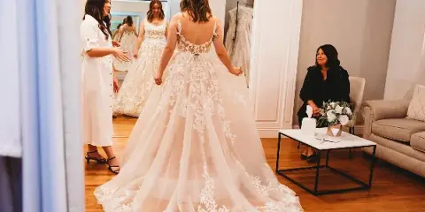 A bride trying on a lace wedding gown in a bridal boutique fitting room with a consultant.