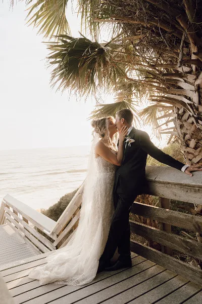 A bride and groom sharing a kiss on a wooden boardwalk near the ocean, framed by palm fronds.