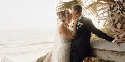 A bride and groom sharing a kiss on a wooden boardwalk near the ocean, framed by palm fronds.