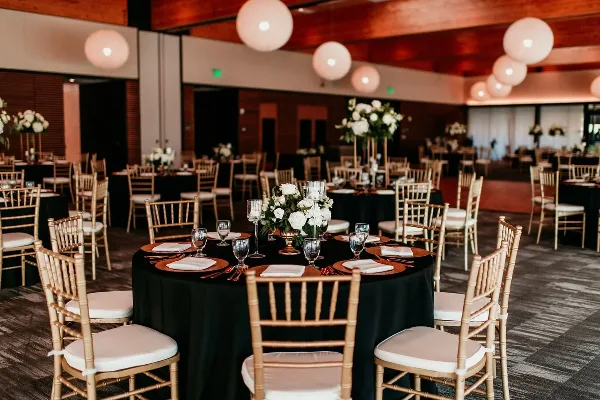 Reception tables set with black linens, gold Chiavari chairs, charger plates, glassware, and floral centerpieces.