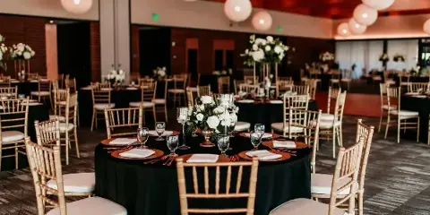 Reception tables set with black linens, gold Chiavari chairs, charger plates, glassware, and floral centerpieces.