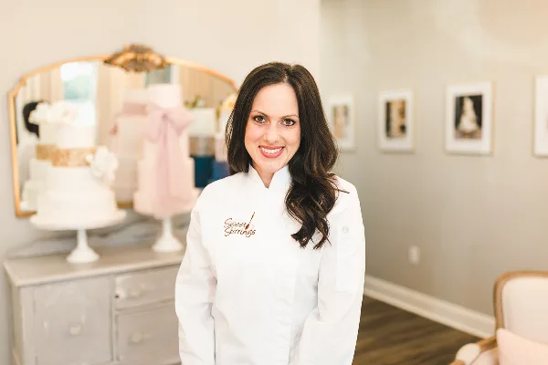 A woman in a white chef's coat with 'Sweet Stirrings' embroidered on the chest stands in a studio with wedding cakes on a counter behind her.
