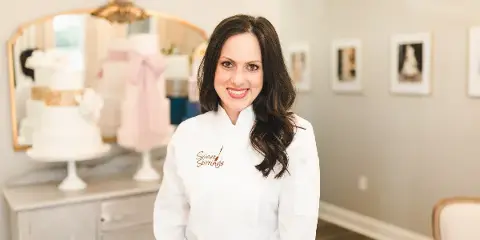 A woman in a white chef's coat with 'Sweet Stirrings' embroidered on the chest stands in a studio with wedding cakes on a counter behind her.