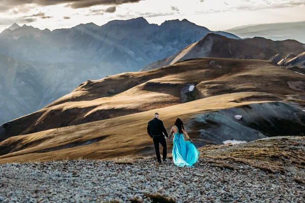 A couple holding hands on a rocky mountain ridge with a vast mountain range in the background.