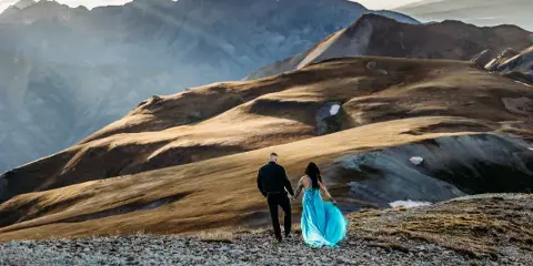 A couple holding hands on a rocky mountain ridge with a vast mountain range in the background.