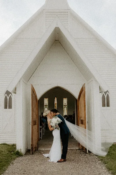 Bride and groom kissing in front of a white chapel with stained glass windows.