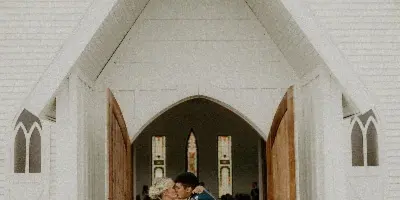 Bride and groom kissing in front of a white chapel with stained glass windows.