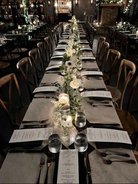 A long wedding reception table featuring a lush floral runner with white roses and greenery, set with menus and glassware in a dimly lit venue.