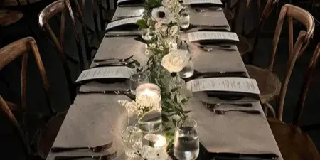 A long wedding reception table featuring a lush floral runner with white roses and greenery, set with menus and glassware in a dimly lit venue.