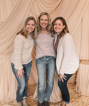 Three smiling women standing together in front of a draped beige fabric backdrop.