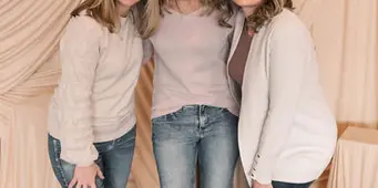 Three smiling women standing together in front of a draped beige fabric backdrop.