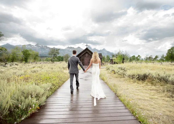 A bride and groom holding hands walking down a wooden boardwalk toward a small chapel with mountains in the background.