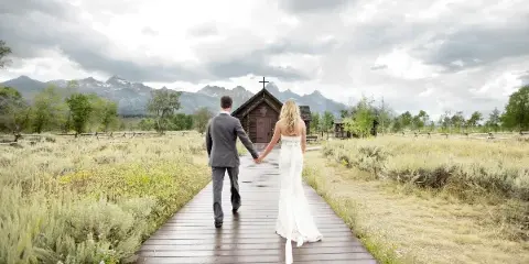 A bride and groom holding hands walking down a wooden boardwalk toward a small chapel with mountains in the background.