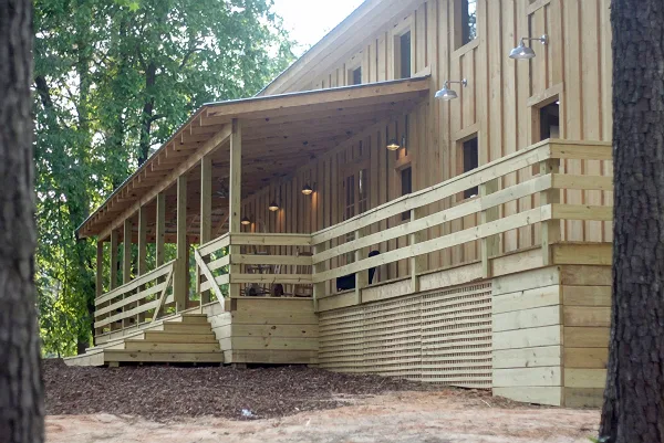 Rustic wooden event venue building with a covered porch, railings, and steps set among trees.