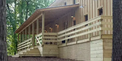 Rustic wooden event venue building with a covered porch, railings, and steps set among trees.