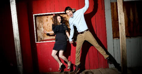 A professional portrait of a man and woman posing playfully against a red wooden wall.