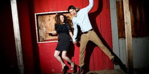 A professional portrait of a man and woman posing playfully against a red wooden wall.