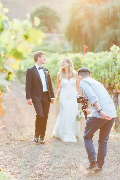 A videographer filming a bride and groom walking through a vineyard.