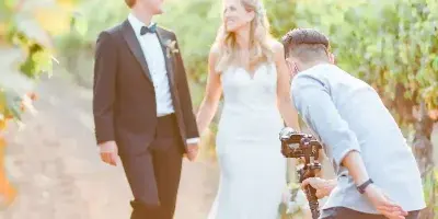 A videographer filming a bride and groom walking through a vineyard.