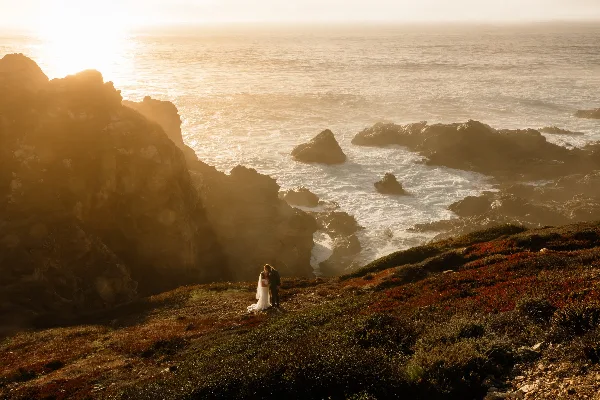 A bride and groom standing on a grassy cliff overlooking the ocean at sunset.