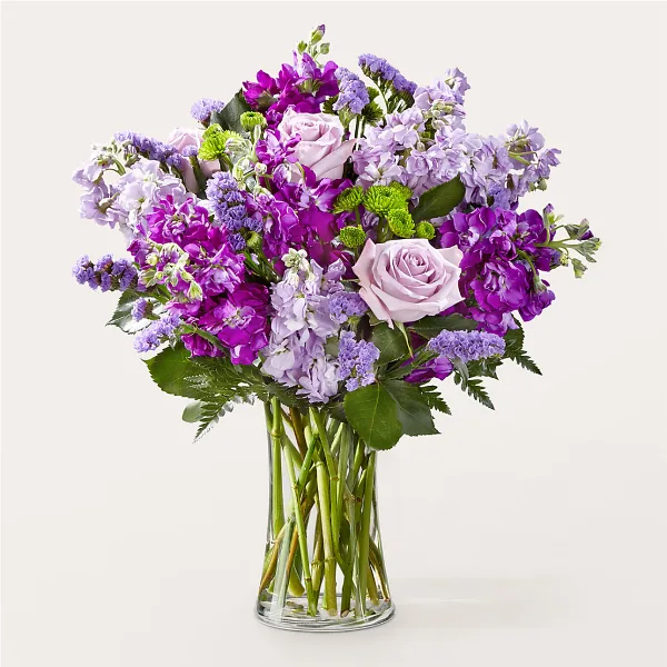 A vibrant floral arrangement featuring purple and pink roses, stock, and greenery in a clear glass vase.