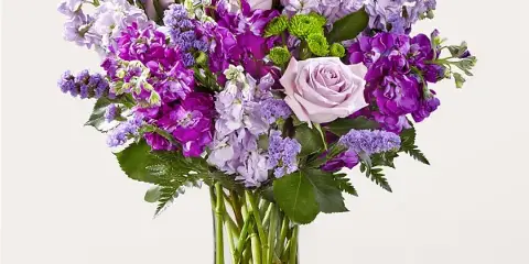 A vibrant floral arrangement featuring purple and pink roses, stock, and greenery in a clear glass vase.