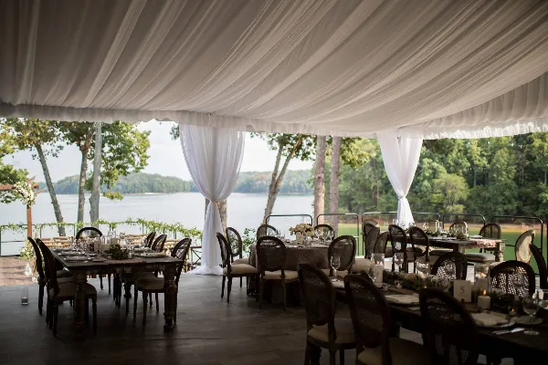 An outdoor wedding reception setup featuring a white draped tent ceiling, set dining tables with glassware, and chairs arranged on a wooden deck overlooking a lake.