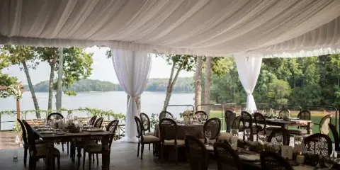 An outdoor wedding reception setup featuring a white draped tent ceiling, set dining tables with glassware, and chairs arranged on a wooden deck overlooking a lake.