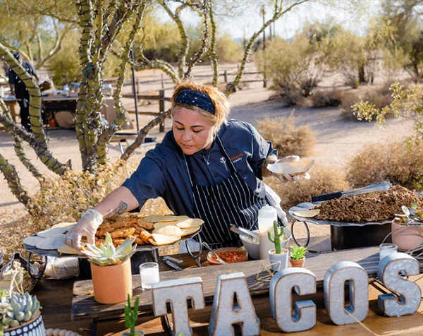 A catering staff member in a dark blue uniform and apron serving food from a buffet table at an outdoor event.