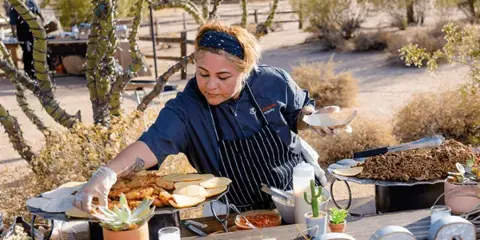 A catering staff member in a dark blue uniform and apron serving food from a buffet table at an outdoor event.