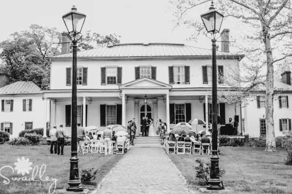 Black and white photo of a wedding ceremony taking place on the front lawn of a historic white Georgian mansion with a portico.